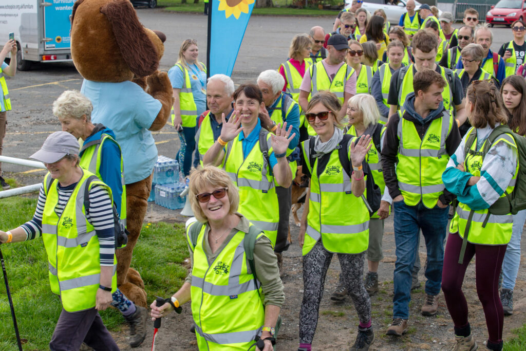 Photo pf Pie and Pint 2023 participants in hi-vis vests smiling and waving as they set off on the 10km walk. Sunny the dog, the Hospice mascot waves them off in the background.