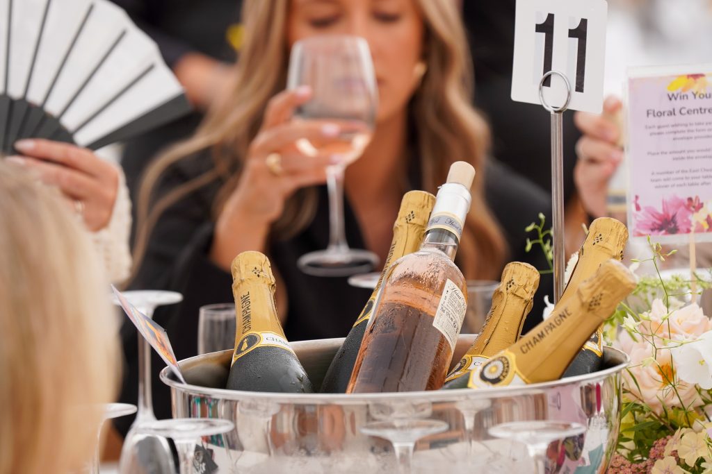 Photo of bottles of wine in a bucket in the centre of a table. A lady is drinking a glass of wine out of focus in the background.