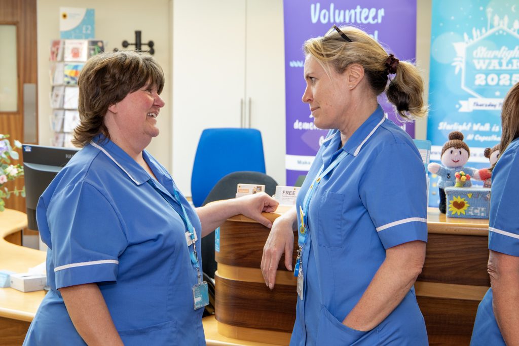 Photo of two Hospice clinical staff in blue uniforms chatting in front of the Hospice reception desk