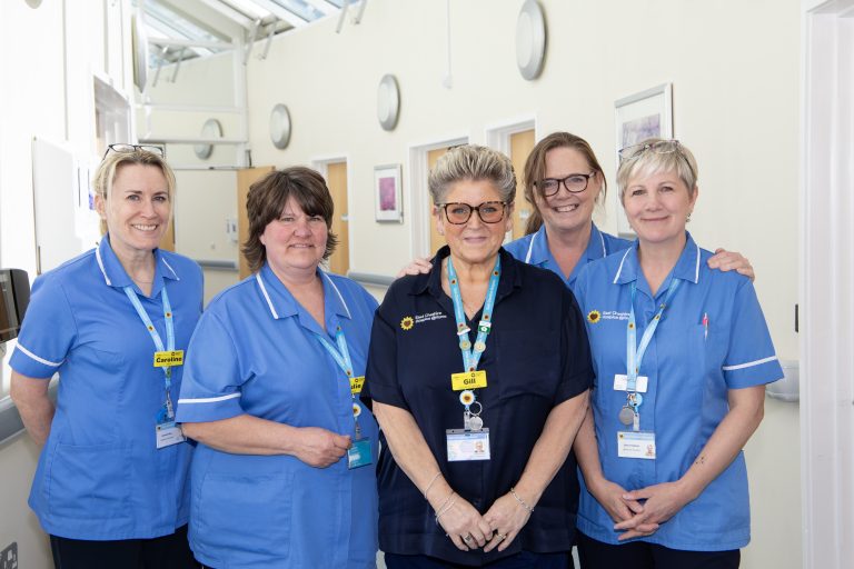 Photo of 5 clinical staff members in blue uniforms in a bright corridor smiling at the camera