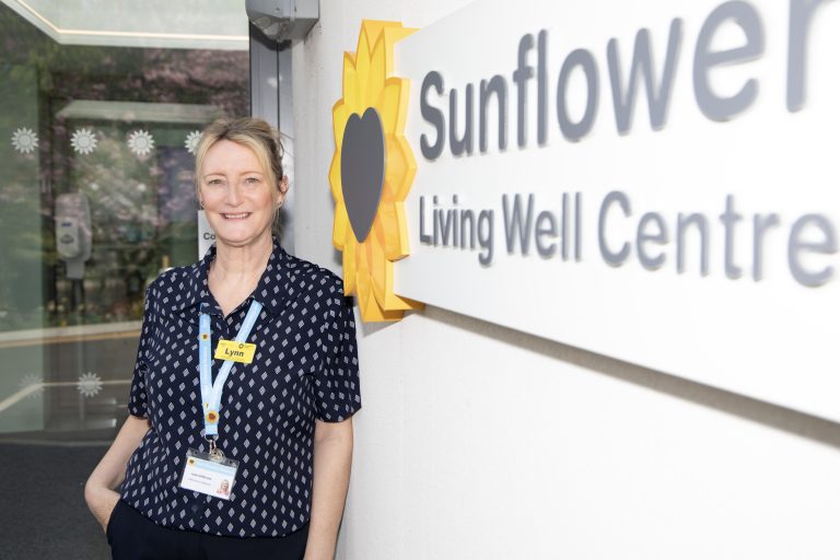 Photo of Sunflower Centre staff member smiling at the camera stood next to a sign that says "Sunflower Living Well Centre".