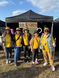 Photo of 5 Starlight Walk participants in green yellow and red bucket hats smiling at the camera in front of a Hair and Glitter stall
