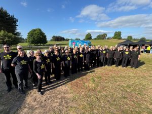 Photo of a group of 40-50 people in black clothing with a yellow star on their t-shirts saying 'ROCK CHOIR' stood in a field smiling at the camera.
