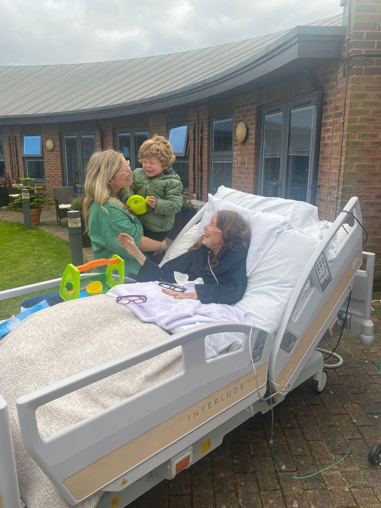 Elaine, in a Hospice bed, with daughter Sophie and grandson Ned by Elaine's side out in the Hospice garden.