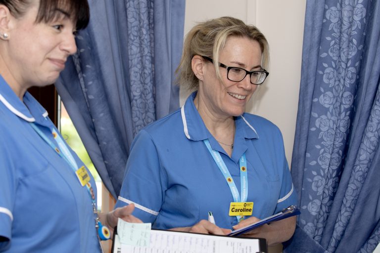 Photo of two Hospice clinical staff with clipboards smiling and chatting to a patient who is out of shot.
