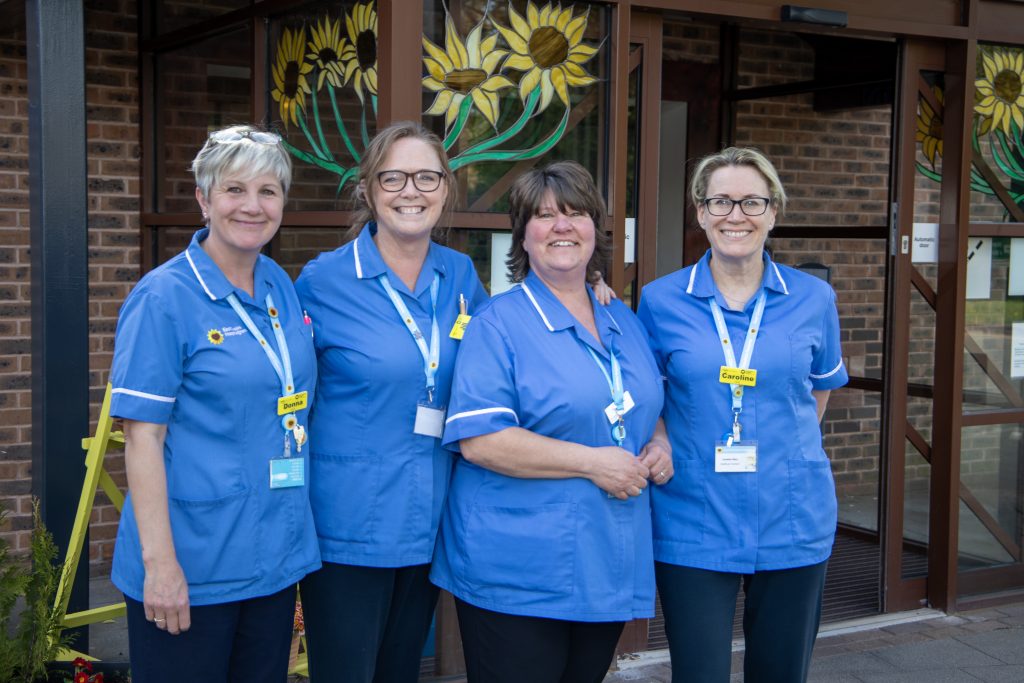 Photo of four clinical staff in blue uniforms stood outside the Hospice entrance smiling at the camera