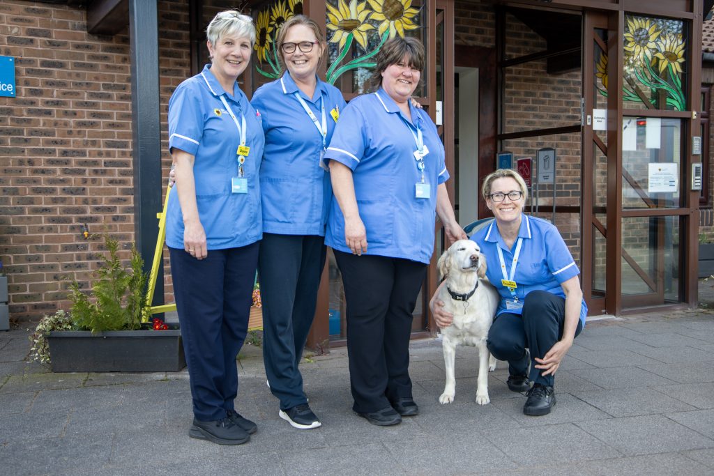 Photo of four clinical staff stood outside the Hospice entrance smiling at the camera with a golden retriever dog between them