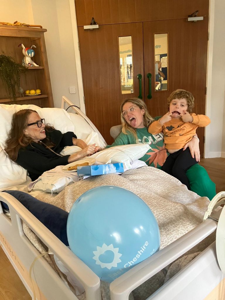 Photo of patient in a Hospice bed having fun with a family members. A lady is sat next to the bed with a child sat on her knee and both are pulling a funny face.