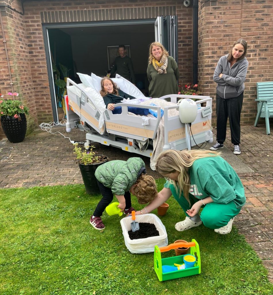 Photo of a patient in a Hospice bed outside in the Hospice garden. They are surrounded by family and they are happily watching a young child doing some gardening.