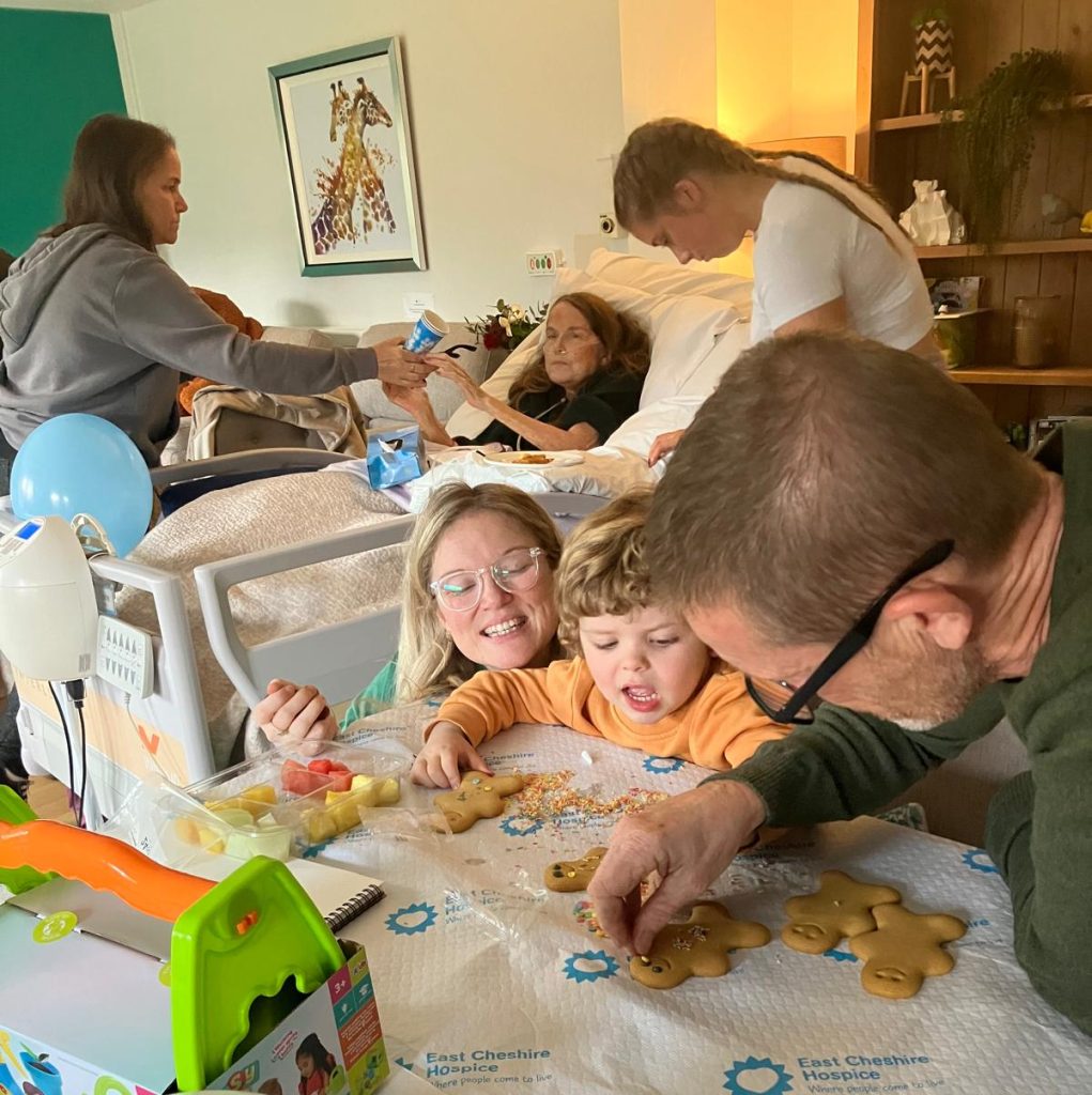 Photo inside the Hospice's family room where a patient is sat up in bed interacting with family members while a young child decorates gingerbread people at a table.