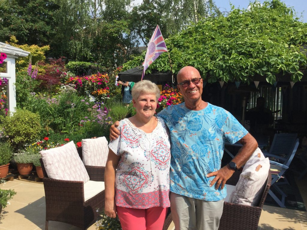 Hospice supporters Julie and Bill North stood smiling at the camera in their garden with flowers and garden furniture in the background.