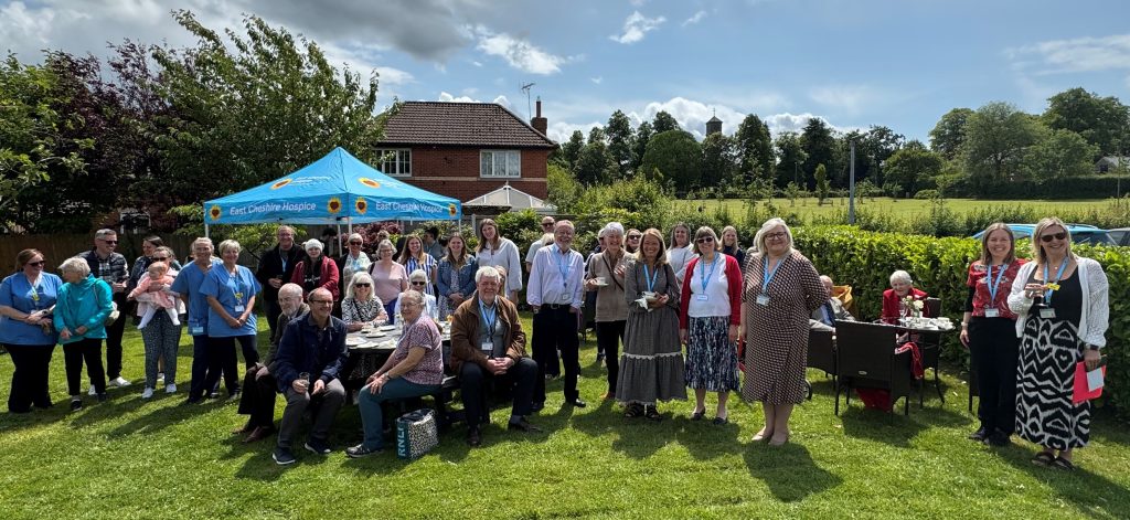 Large group photo of memory tree party attendees, Hospice staff and Trustees, all looking at the camera on a sunny day.