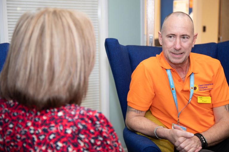 Photo of a Dementia Services team member in an orange polo shirt sat talking to another individual who is facing away from the camera.