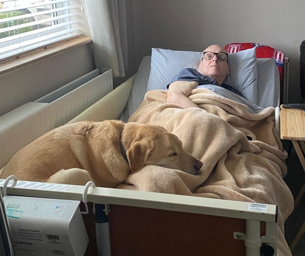 Photo of Hospice patient Shane in bed with pet Labrador lying at the bottom of the bed