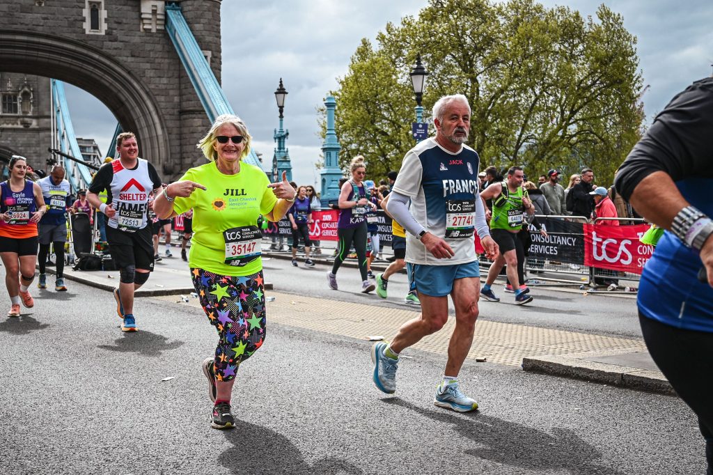 Jill Booth running on London Bridge in one of her races pointing at her bright yellow Hospice t-shirt.