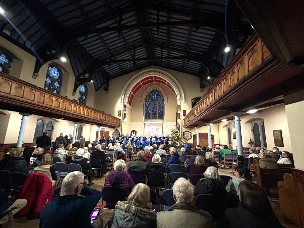 Photo from the back of a church looking towards the front during a service. Backs of the heads of the congregation are visible in the foreground with a choir performing in the background.