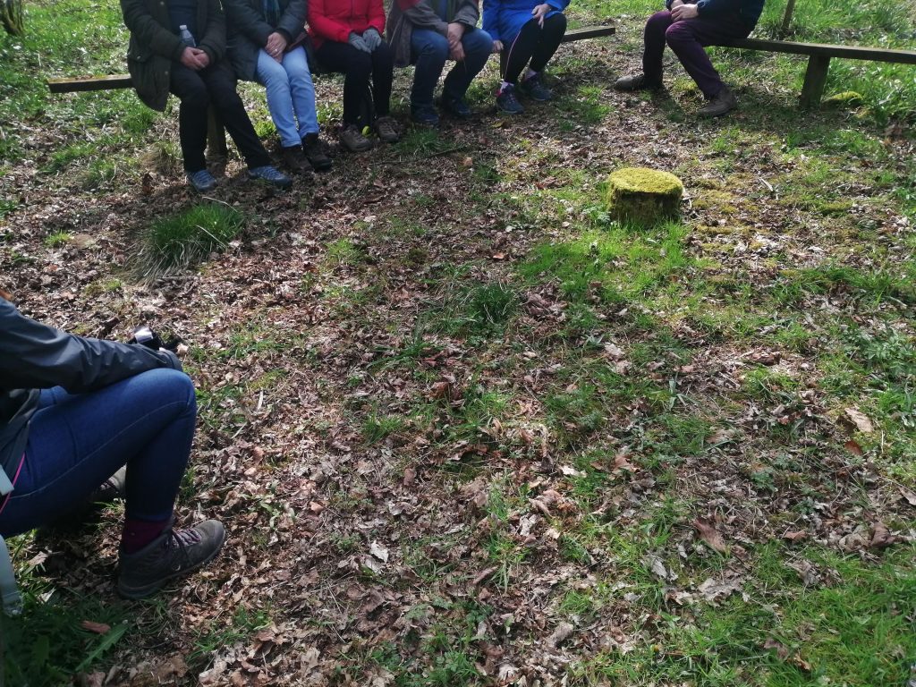 Photo of people sat together in a forest clearing. Only their lower halves are in the shot.