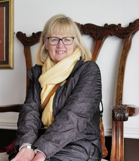 Photo of a lady sat in a large wooden chair wearing a light coloured scarf and a dark jacket smiling at the camera