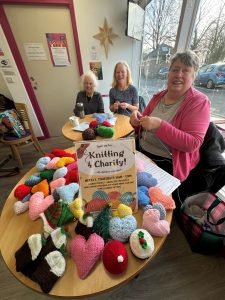 Photo of three people sat around tables in a cafe with a large pile of knitted items. A sign on the table says "knitting &=4 Charity!".