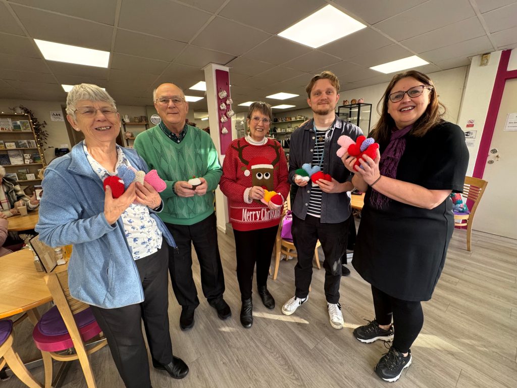 Photo of five people stood in a café posing with knitted items.