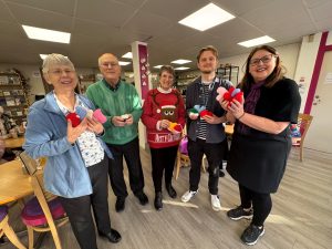 Photo of five people stood in a café posing with knitted items.