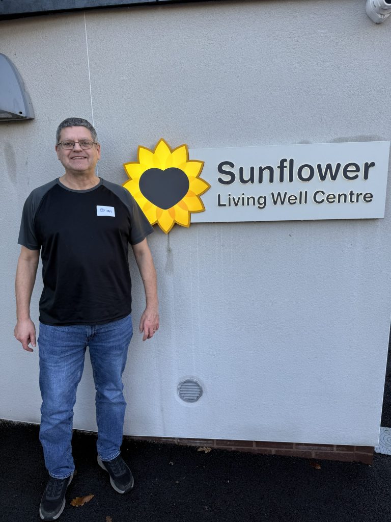 Photo of a man in a black t-shirt and jeans stood outside the Hospice's Sunflower Living Well Centre next o its illuminated sign.