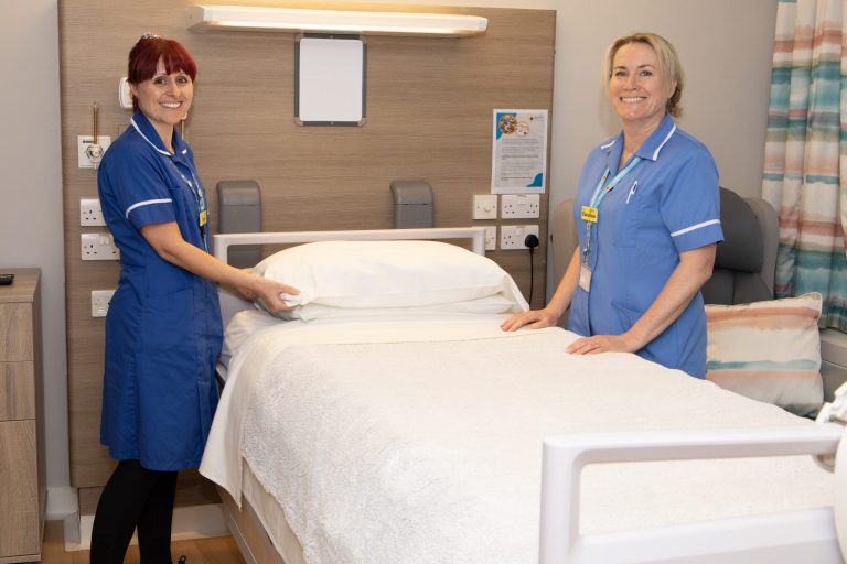 Photo of two nurses in uniform stood either side of a Hospice bed smiling warmly at the camera