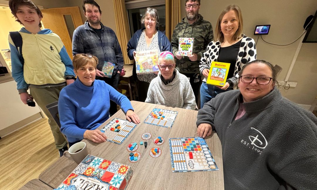 Group of 8 people stood smiling at the camera around a table with board games on.