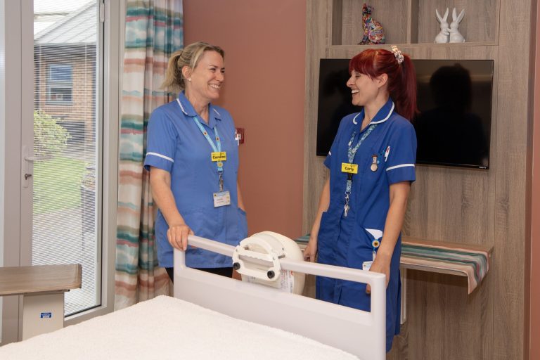 Photo of two clinical staff members in uniform stood at the end of the bed in an inpatient room laughing with each other