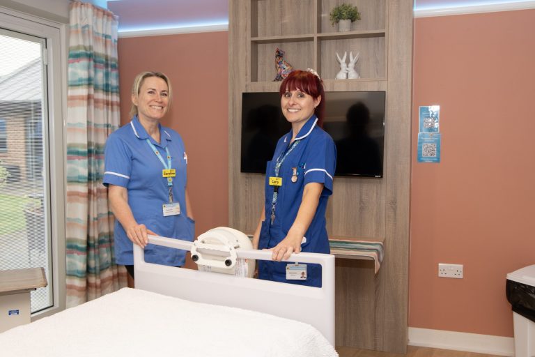 Photo of two nurses in uniform stood at the end of a Hospice bed smiling warmly at the camera