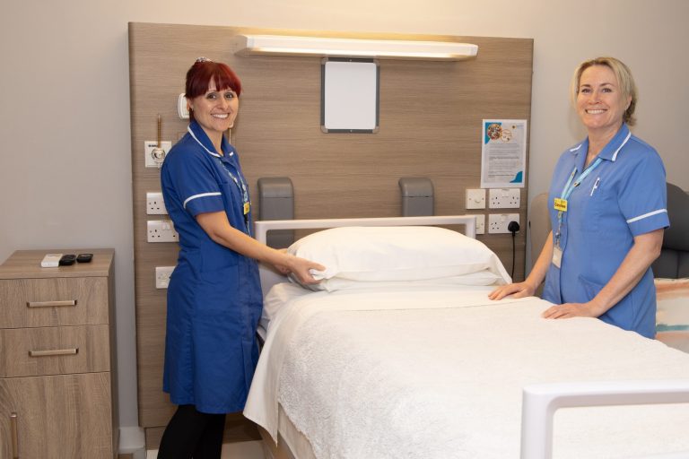 Photo of two nurses in uniform stood either side of a Hospice bed smiling warmly at the camera