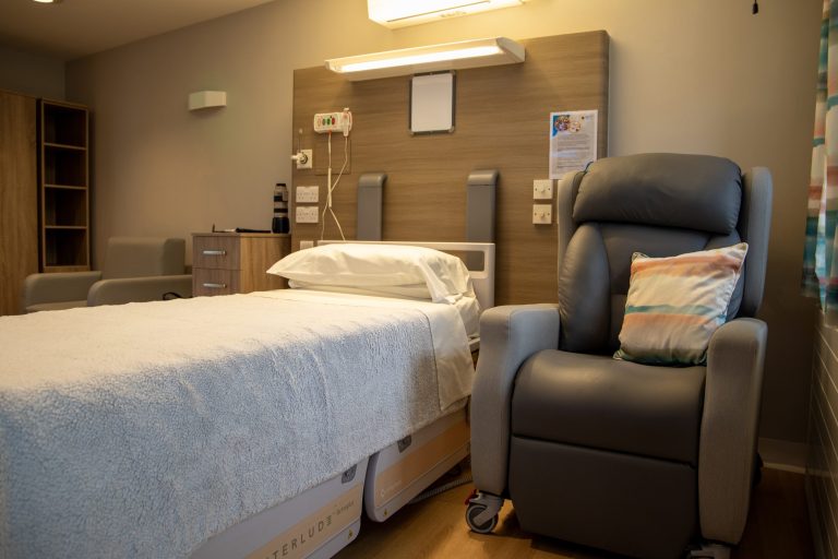 Photo of a private bedroom in the Hospice inpatient unit featuring a neatly made bed and comfy arm chair.