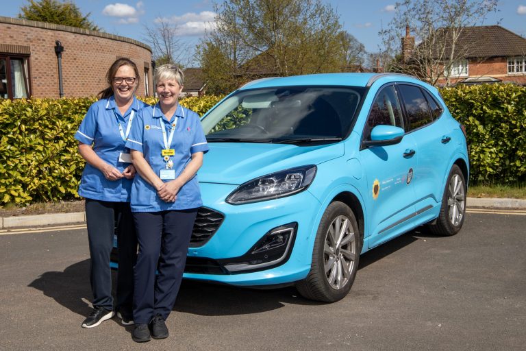 Photo of two nurses stood in front of the Hospice @Home car smiling at the camera on a sunny day