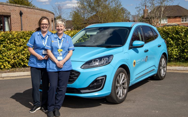 Photo of two nurses stood in front of the Hospice @Home car smiling at the camera on a sunny day