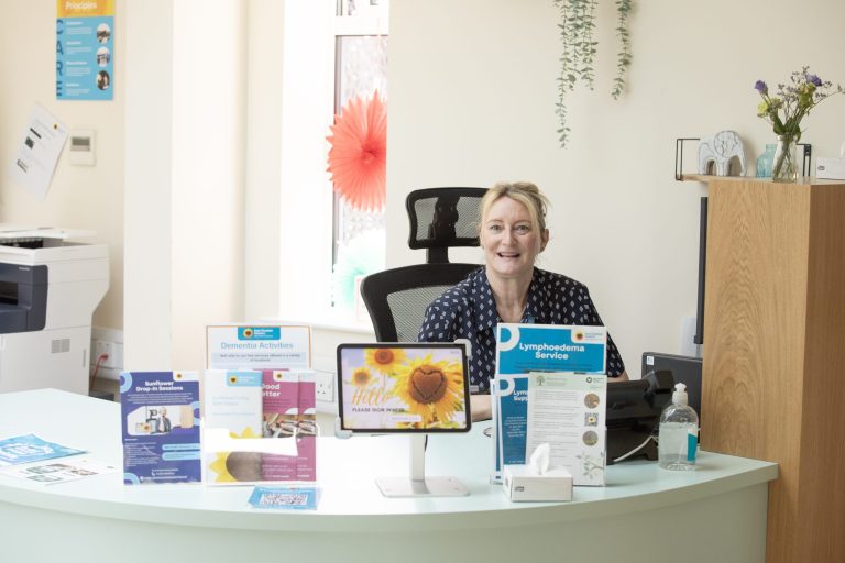 Photo of a lady sat behind the reception desk in the Sunflower Living Well Centre on a sunny day.