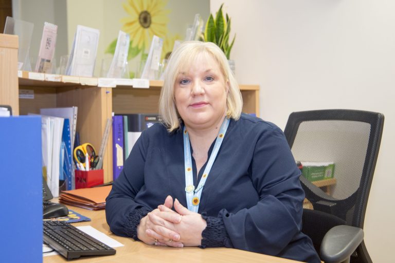 A lady who works with the Hospice is sitting at her desk smiling at the camera