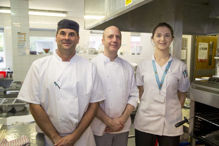 Three people are stood in a kitchen smiling