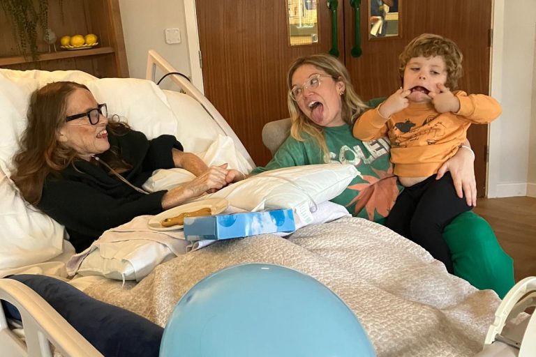 Photo of patient in a Hospice bed having fun with a family members. A lady is sat next to the bed with a child sat on her knee and both are pulling a funny face.