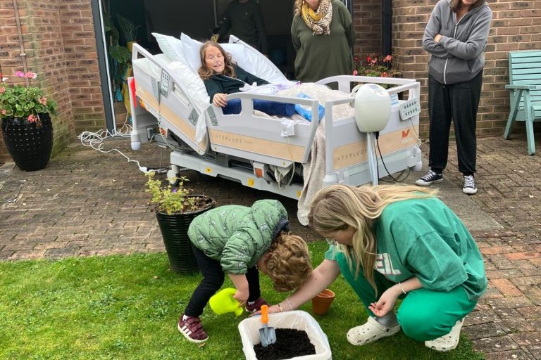 Photo of a patient in a Hospice bed outside in the Hospice garden. They are surrounded by family and they are happily watching a young child doing some gardening.