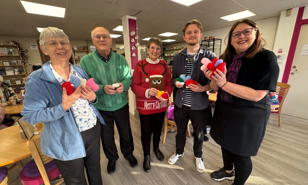 Photo of five people stood in a café posing with knitted items.