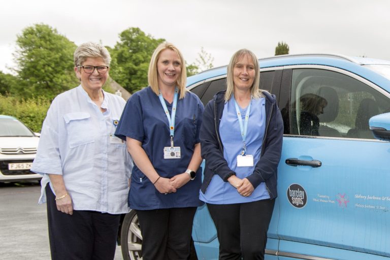 Three Hospice @Home staff next to a blue Hospice @Home smiling at the camera