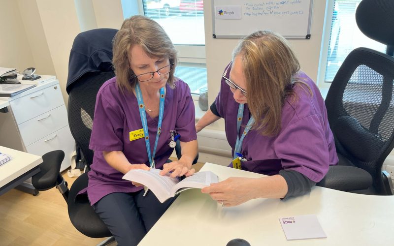 Photo of two PACE staff in purple uniforms looking at a notebook together in a bright office.
