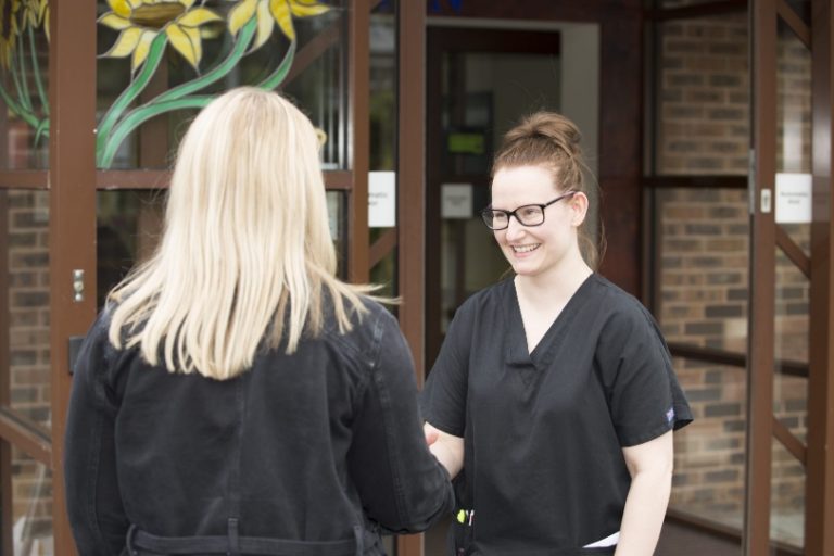 Two people shaking hands outside the Hospice entrance, the back of one person's head is visible and the other is facing the first person, smiling, wearing a black clinical uniform and glasses.