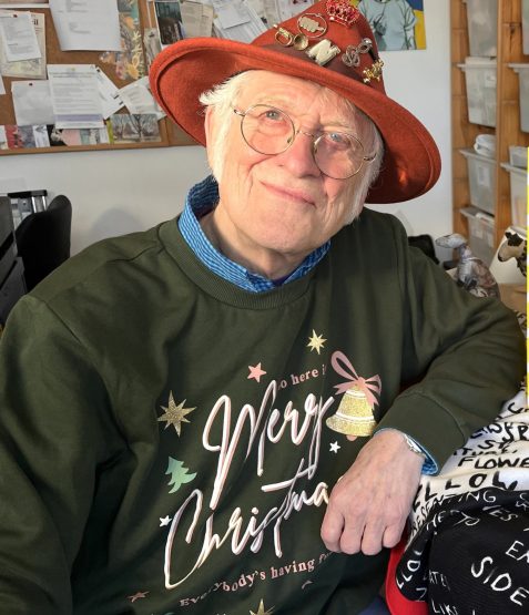 Photo of Noddy Holder in colourful clothing sat smiling at the camera