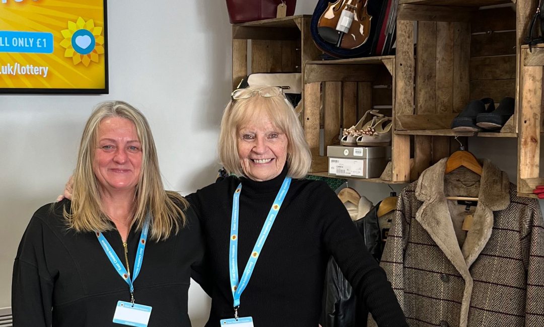Two volunteers stood smiling at the camera stood behind the reception desk at East Cheshire Hospice's Congleton shop