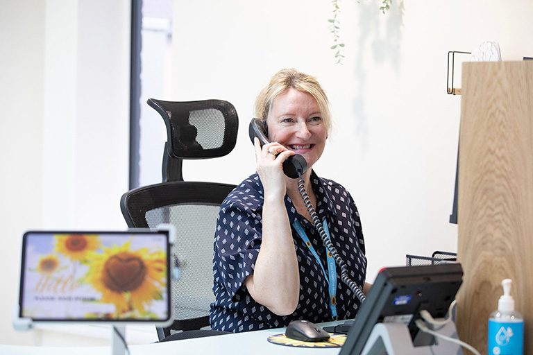 A lady who works at East Cheshire Hospice is sat down at her desk smiling whilst on the phone