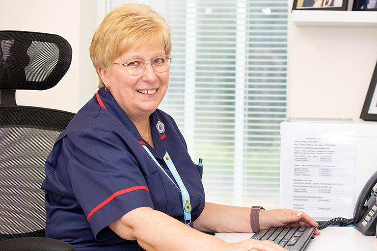 A lady who works at East Cheshire Hospice sat down at her desk with her nurse uniform on smiling at the camera