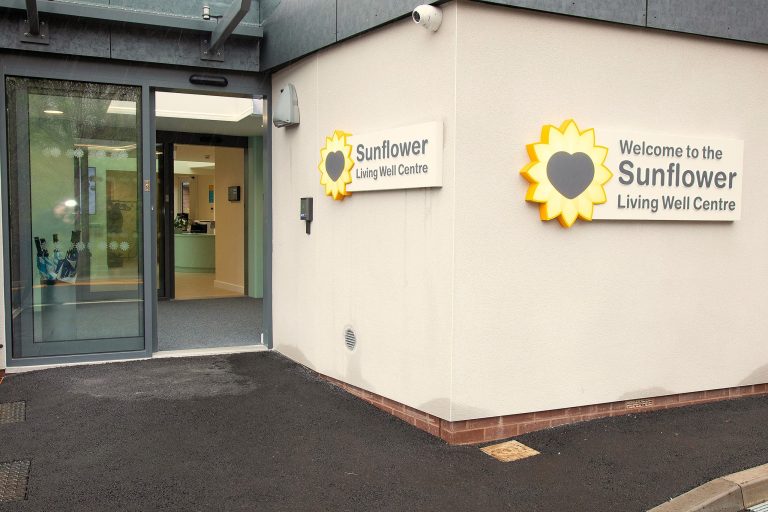 Photo of the entrance of the Sunflower Living Well Centre consisting of sliding glass doors on the left and a light rendered wall on the right with an illuminated Sunflower Centre sign and Hospice logo.